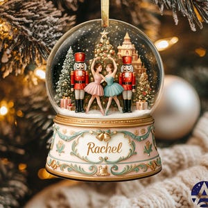 May include: A snow globe with a ballet scene, including two dancers, two nutcracker soldiers, and a Christmas tree. The pink and gold base has the name "Rachel". The globe hangs from a gold ribbon. A festive Christmas ornament.