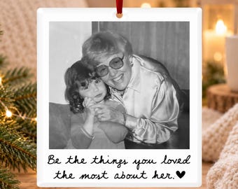 May include: A square white ceramic ornament featuring a black and white photograph of an adult and a child smiling together. The text below the photo reads, "Be the things you loved the most about her." with a small black heart symbol at the end.