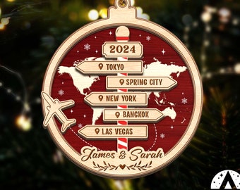 May include: A round wooden Christmas ornament with a red and white striped candy cane design. The ornament features a world map with a plane and five destination signs that read "2024", "Tokyo", "Spring City", "New York", "Bangkok", and "Las Vegas". The ornament is personalized with the names "James & Sarah".
