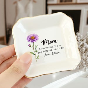 May include: A square, off-white ceramic trinket dish with a gold rim. The dish features a purple flower illustration and the text "Mom, Everything I am you helped me to be Love, Olivia."
