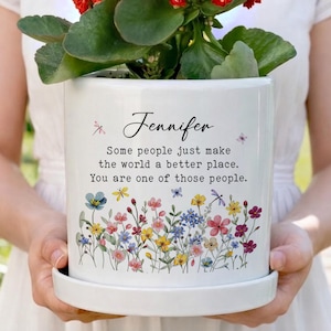 May include: White ceramic plant pot with a floral design and the name "Jennifer" printed on it. The pot contains a red flowering plant. The text on the pot reads "Some people just make the world a better place. You are one of those people."
