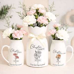 May include: Three white ceramic vases with floral arrangements. The vases are decorated with text and floral illustrations. The center vase has a bow. The text on the vases reads "Chapter Forty", "Chapter Thirty", and "Chapter Twenty-One". The vases contain pink and white flowers.