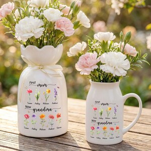 Pu&ograve; includere: Due vasi in ceramica bianca, uno alto e uno a forma di tazza, riempiti di garofani bianchi e rosa. I vasi sono decorati con illustrazioni floreali e il testo "First Mom Now Grandma" con nomi sotto.