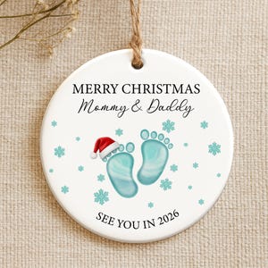 May include: White ceramic Christmas ornament with the text "MERRY CHRISTMAS Mommy & Daddy" and "SEE YOU IN 2026". Features baby footprints, a Santa hat, and snowflakes. Includes a jute twine hanger.