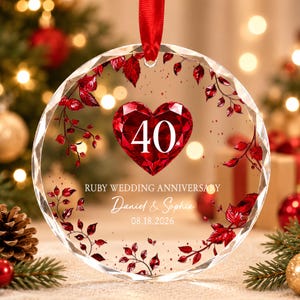Op de afbeelding: Een rond ornament van helder glas met een gefacetteerd rood hart met het getal 40 in het wit. De tekst luidt RUBY WEDDING ANNIVERSARY, Daniel & Sophia en 08.18.2026. Rode bladeren versieren de randen en er is een rood lint bevestigd.