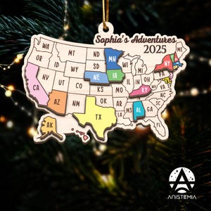 May include: Wooden Christmas ornament in the shape of the United States, with coloured states. The ornament reads "Sophia's Adventures 2025" and hangs from a gold string. The ornament is made of light-coloured wood. States are highlighted in various colours.