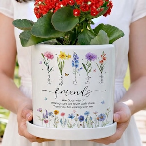 May include: White ceramic planter with colorful floral illustrations and the word "friends" in script. The planter features names and a quote: "Are God's way of making sure we never walk alone. Thank you for walking with me." A red flowering plant sits inside.