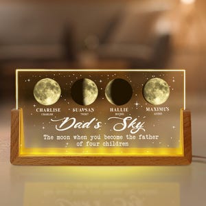 May include: A personalized wooden night light featuring four moon phase images with names and dates. The text "Dad's Sky" and a message are displayed below the moon phases. The light has a warm glow.
