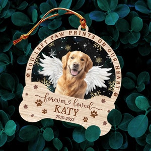 May include: A wooden Christmas ornament in the shape of a bone with a golden retriever dog with angel wings inside a circle. The text on the ornament reads "You left paw prints on our heart forever loved KATY 2010-2023".