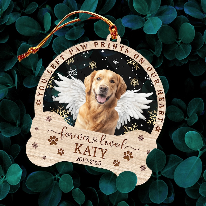 May include: A wooden Christmas ornament in the shape of a bone with a golden retriever dog with angel wings inside a circle. The text on the ornament reads "You left paw prints on our heart forever loved KATY 2010-2023".