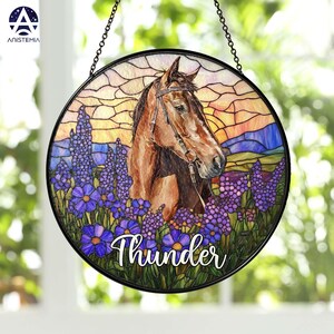 Pode incluir: Um apanhador de sol circular de vitral apresentando um cavalo castanho com r&eacute;deas, rodeado por flores roxas num campo. O fundo mostra um c&eacute;u ao p&ocirc;r do sol com tons de laranja e amarelo. A palavra Thunder est&aacute; escrita em branco.