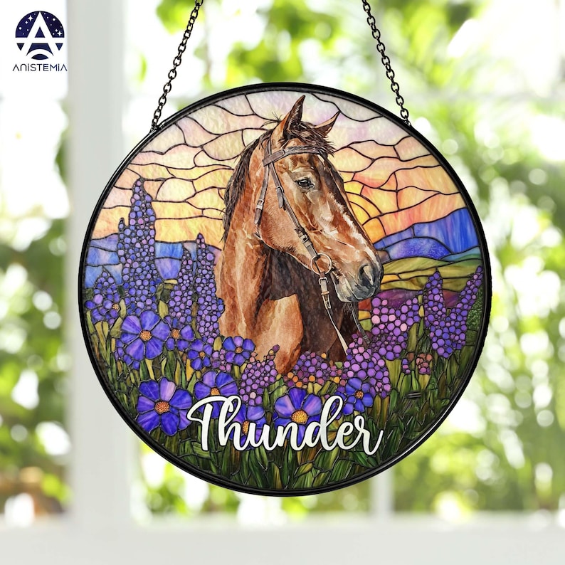 Pode incluir: Um apanhador de sol circular de vitral apresentando um cavalo castanho com r&eacute;deas, rodeado por flores roxas num campo. O fundo mostra um c&eacute;u ao p&ocirc;r do sol com tons de laranja e amarelo. A palavra Thunder est&aacute; escrita em branco.
