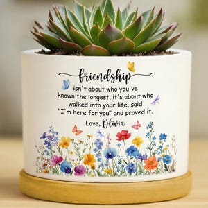 May include: A white ceramic cylindrical planter with a wooden base containing a green succulent. The planter features a colourful wildflower print, butterflies, and the text: friendship isn't about who you've known the longest, it's about who walked into your life, said "I'm here for you" and proved it. Love, Olivia.