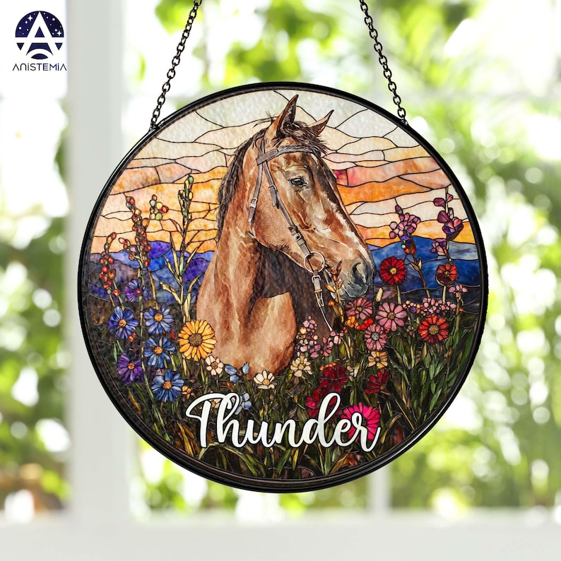 Pode incluir: Um apanhador de sol circular em vitral com um cavalo castanho com r&eacute;deas, rodeado de flores silvestres coloridas e uma paisagem ao p&ocirc;r do sol. A palavra Thunder est&aacute; escrita em letra branca na parte inferior da moldura.