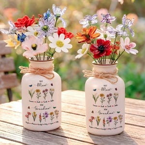 以下が含まれることがあります： カラフルな造花が入ったクリーム色の花瓶が2つ。各花瓶にはロープのアクセントがあり、「First Mom Now Grandma」の文字と名前と花のイラストが描かれています。花には赤、白、オレンジ、紫の花が含まれています。