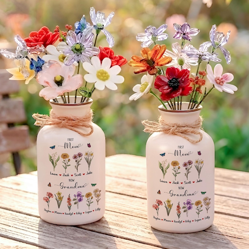 以下が含まれることがあります： カラフルな造花が入ったクリーム色の花瓶が2つ。各花瓶にはロープのアクセントがあり、「First Mom Now Grandma」の文字と名前と花のイラストが描かれています。花には赤、白、オレンジ、紫の花が含まれています。