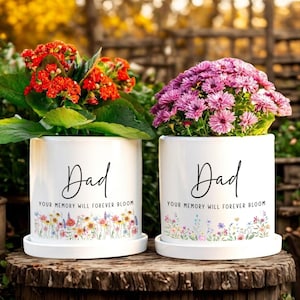 May include: Two white ceramic flower pots with the word "Dad" in black script. The pots are filled with colorful flowers and have the text "YOUR MEMORY WILL FOREVER BLOOM". The pots sit on a wooden surface.