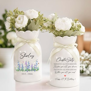 May include: Two white ceramic vases with white flowers and green foliage. One vase says "Shirley" with "EST. 1966" and a floral design. The other vase says "Chapter Sixty" with a birthday message. Both vases have cream-colored ribbon.