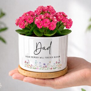 May include: A white ceramic planter with the word "Dad" in black script, filled with vibrant pink flowers. The planter has a floral design at the base and sits on a wooden coaster. The text "YOUR MEMORY WILL FOREVER BLOOM" is also on the planter.