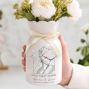 May include: White ceramic vase with a map design in the shape of a house, tied with a cream ribbon. The vase holds white flowers and greenery. Text on the vase reads "OUR FIRST HOME" and the names "Amelia & Lucas".
