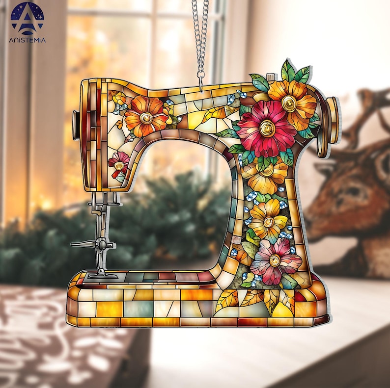 May include: A decorative sewing machine ornament with a stained-glass effect. The ornament features a colorful floral design with red, orange, and yellow flowers. The sewing machine is suspended by a silver chain. The word "ANISTEMIA" is printed on the top left.