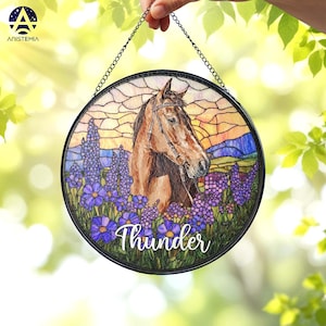 Pode incluir: Um apanhador de sol circular em vitral com um cavalo castanho com r&eacute;deas, rodeado por flores roxas contra um c&eacute;u ao p&ocirc;r do sol. O nome Thunder est&aacute; escrito em letras brancas na parte inferior. Pendurado por uma corrente de metal preta.