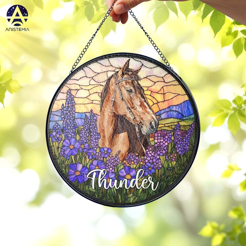 Pode incluir: Um apanhador de sol circular em vitral com um cavalo castanho com r&eacute;deas, rodeado por flores roxas contra um c&eacute;u ao p&ocirc;r do sol. O nome Thunder est&aacute; escrito em letras brancas na parte inferior. Pendurado por uma corrente de metal preta.