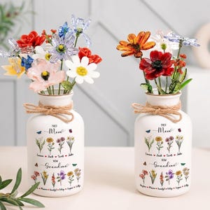 以下が含まれることがあります： 白いセラミックの花瓶が2つ、生け花が飾られています。各花瓶には紐のリボンと「First Mom Now Grandma」の文字、名前と花のイラストが描かれています。花は赤、オレンジ、黄色、ピンク、白の色合いです。