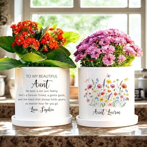 May include: Two white ceramic flower pots with saucers, each holding vibrant flowers. One pot has red flowers and text that reads "To My Beautiful Aunt." The other pot has purple flowers and a floral design with the name "Aunt Lauren."