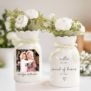 May include: Two white ceramic vases with scalloped rims, each tied with a light cream ribbon. One vase features a printed photograph of two children and the text "Evelyn & Mateo". The other vase displays the text "sister EST. 1997", "maid of honor EST. 2026", and a small heart icon.