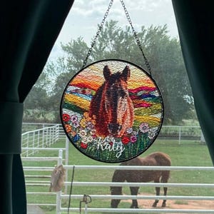 Pode incluir: Um apanhador de sol circular em vitral com uma cabe&ccedil;a de cavalo castanha centrada entre flores coloridas e uma paisagem de p&ocirc;r do sol. O nome Katy est&aacute; escrito em letras brancas na parte inferior. Suspenso por uma corrente de metal.