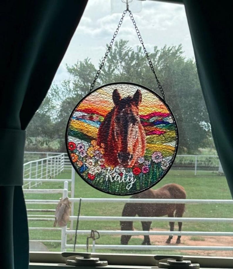 Pode incluir: Um apanhador de sol circular em vitral com uma cabe&ccedil;a de cavalo castanha centrada entre flores coloridas e uma paisagem de p&ocirc;r do sol. O nome Katy est&aacute; escrito em letras brancas na parte inferior. Suspenso por uma corrente de metal.