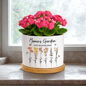May include: A white ceramic flower pot with the text "Mama's Garden LOVE BLOOMS HERE" and names below colorful flower illustrations. The pot holds a vibrant pink flowering plant and sits on a round wooden coaster.