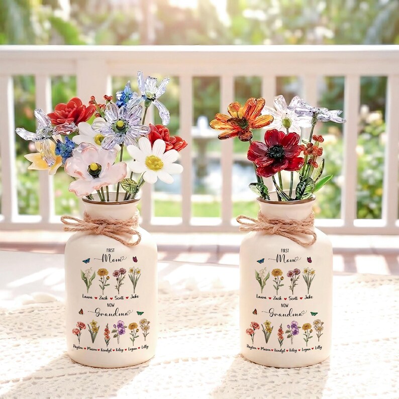以下が含まれることがあります： クリーム色の花瓶2つ。それぞれに花の配置があり、ひもでできたリボンと「First Mom Now Grandma」の文字、名前と花のイラストが描かれています。花はカラフルでガラス製です。