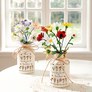 以下が含まれることがあります： 紐で結ばれた、カラフルな造花が入った2つのクリーム色の花瓶。花瓶には「First Mom Now Grandma」の文字と花のイラストが描かれています。花には赤、白、黄色、紫の花が含まれています。