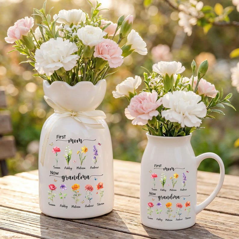 Pu&ograve; includere: Due vasi in ceramica bianca, uno alto e uno a forma di tazza, riempiti di garofani bianchi e rosa. I vasi sono decorati con illustrazioni floreali e il testo "First Mom Now Grandma" con nomi sotto.