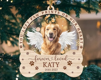 May include: A wooden Christmas ornament shaped like a bone with a golden retriever dog inside. The ornament has the text "You left paw prints on our heart" and "forever loved KATY 2010-2023".
