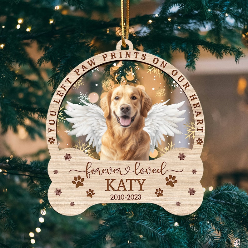 May include: A wooden Christmas ornament shaped like a bone with a golden retriever dog inside. The ornament has the text "You left paw prints on our heart" and "forever loved KATY 2010-2023".