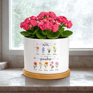 May include: A white ceramic planter on a circular wooden base containing a vibrant pink Kalanchoe plant. The planter features a floral graphic with the text First Mom Now Grandma and names including Robert, Jessica, Sophia, Shirley, Frank, Diane, and Natalie.