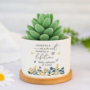 May include: A white ceramic planter with a faux succulent. The planter has the text "carried for a moment, loved for a lifetime" with a name and date. The base is a round wooden coaster. The planter is decorated with blue and yellow flowers.