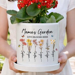 May include: White ceramic planter with the text "Mama's Garden LOVE BLOOMS HERE" and names below floral illustrations. The planter holds a red flowering plant. Includes a matching saucer and butterfly accents.