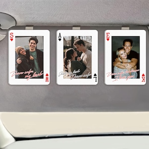 May include: Three white playing card-shaped car visor clips with photos of couples. Each card has a different suit symbol and a handwritten message in red. The cards are attached to a gray car visor.