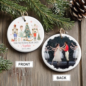May include: A round Christmas ornament with a front and back design. The front features a Nutcracker theme with text that reads "THE NUTCRACKER 2025" and "Sophia AS CLARA". The back shows a ballet scene. A string is attached for hanging.