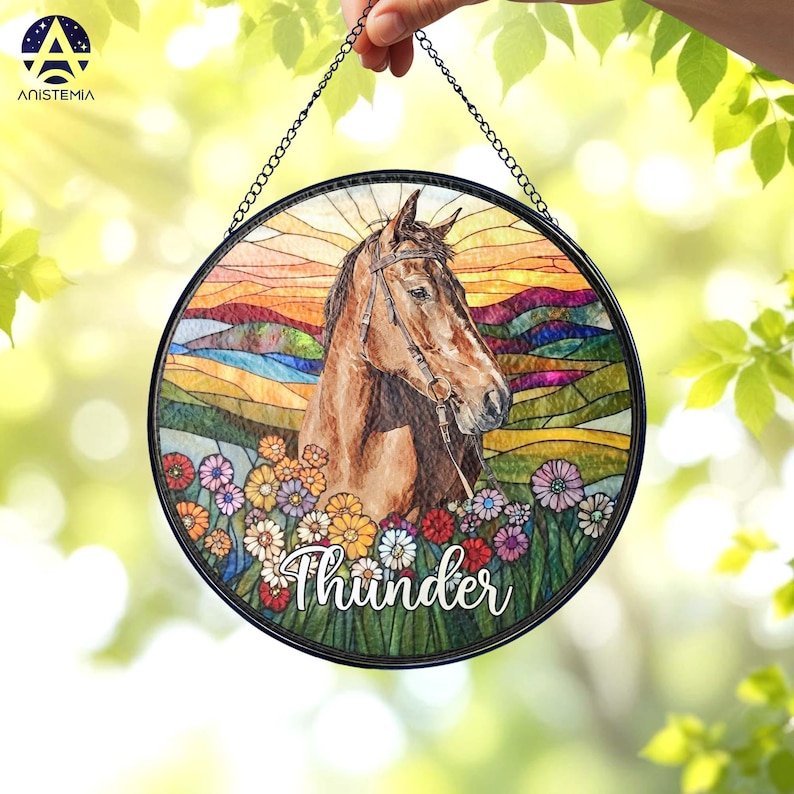 Pode incluir: Um apanhador de sol circular em vitral com um cavalo castanho com r&eacute;deas, sobre uma paisagem colorida de colinas e um c&eacute;u ao p&ocirc;r do sol. O primeiro plano mostra um campo de flores multicoloridas com o nome Thunder escrito em branco.
