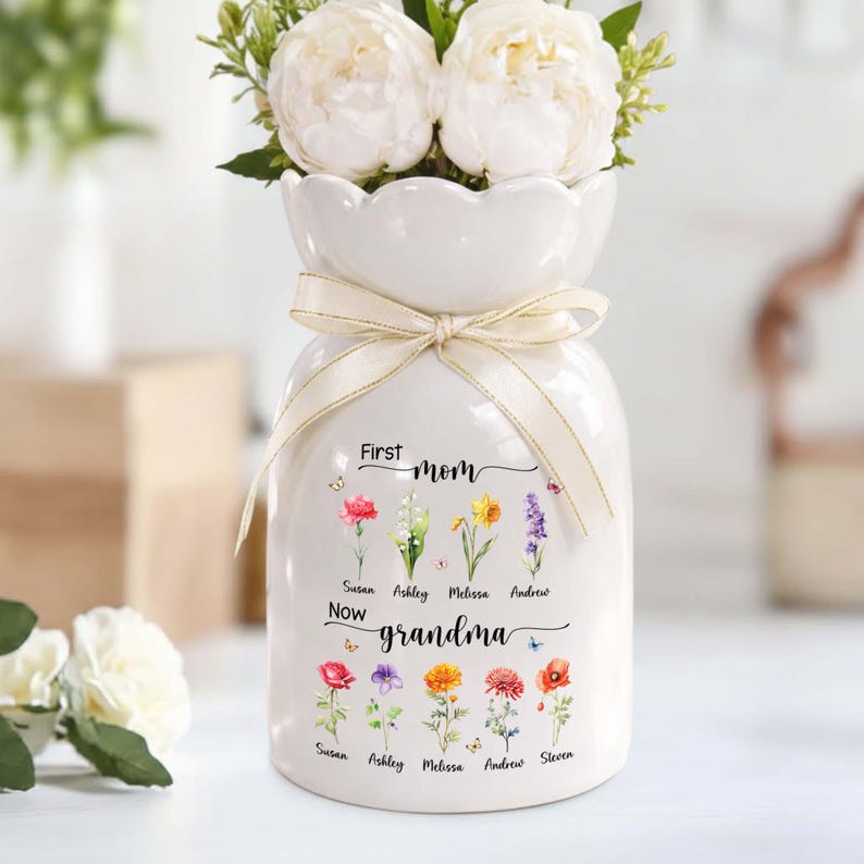 Pu&ograve; includere: Un vaso di ceramica bianco con un nastro color crema legato attorno al collo. Il vaso &egrave; decorato con illustrazioni floreali colorate e le parole "First Mom Now Grandma". Il vaso contiene fiori bianchi.
