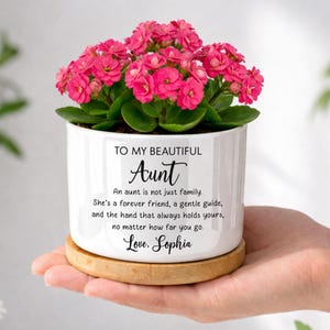 May include: A white ceramic planter with a bamboo base containing a vibrant pink Kalanchoe plant. The planter features black printed text that reads: TO MY BEAUTIFUL Aunt, An aunt is not just family. She's a forever friend, a gentle guide, and the hand that always holds yours, no matter how far you go. Love, Sophia.