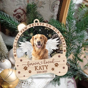 May include: A wooden Christmas ornament shaped like a bone with a photo of a golden retriever with angel wings inside a circle. The text "You left paw prints on our heart" is written around the circle. The text "forever loved KATY 2010-2023" is written below the circle.