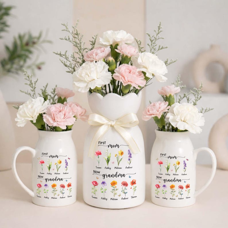 Pu&ograve; includere: Tre vasi in ceramica bianca con composizioni floreali. I vasi sono decorati con le parole "First Mom Now Grandma" e illustrazioni floreali. I fiori sono garofani bianchi e rosa. Il vaso centrale ha un fiocco.