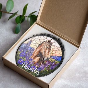 Pode incluir: Um apanhador de sol circular em vitral com um cavalo castanho num campo de flores de tremo&ccedil;o roxas. A pe&ccedil;a est&aacute; emoldurada em metal preto com uma corrente e apresenta o nome Thunder em escrita branca dentro de uma caixa de presente.