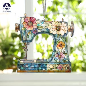 May include: A decorative sewing machine ornament with a stained glass effect. The ornament features a floral design with pink, yellow, and purple flowers. The sewing machine is blue and has a gold-colored handle and needle.